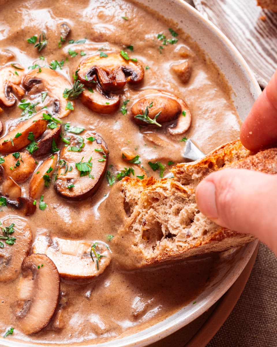 Crusty bread being dunked into a bowl of chunky vegan mushroom soup.