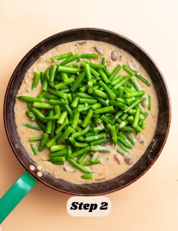 The second step in the process of making green bean casserole: the image shows the cooked green beans being transferred to a creamy mushroom sauce in a large skillet.