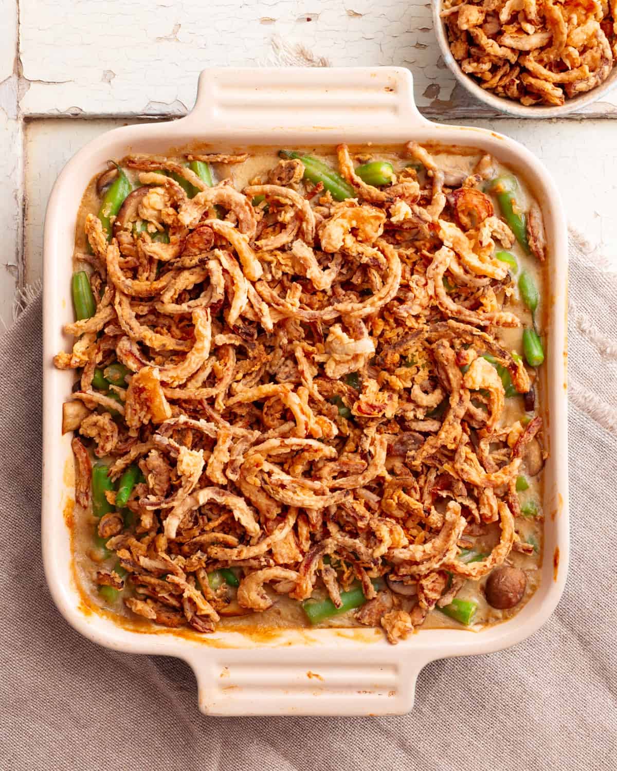 A top-down view of a vegan green bean casserole, topped with crispy golden brown onions. The casserole is in a ceramic dish on top of a weathered, white wooden table. 