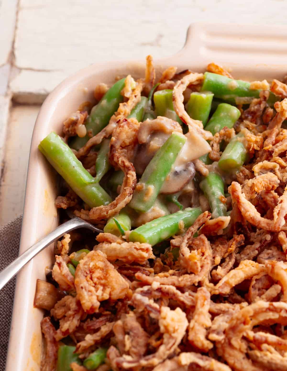 An antique silver spoon taking a scoop of vegan green bean casserole from a ceramic dish. 