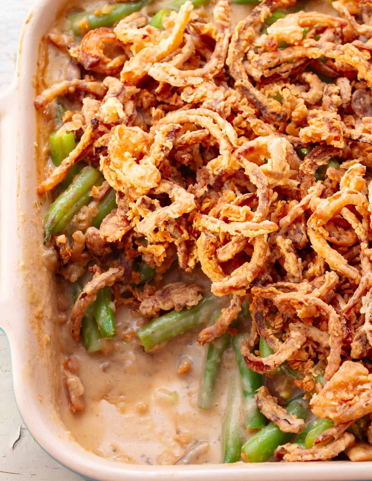 a ceramic dish of vegan green bean casserole with a portion taken out. The creamy sauce and the green beans are visible under a layer of crispy onions.