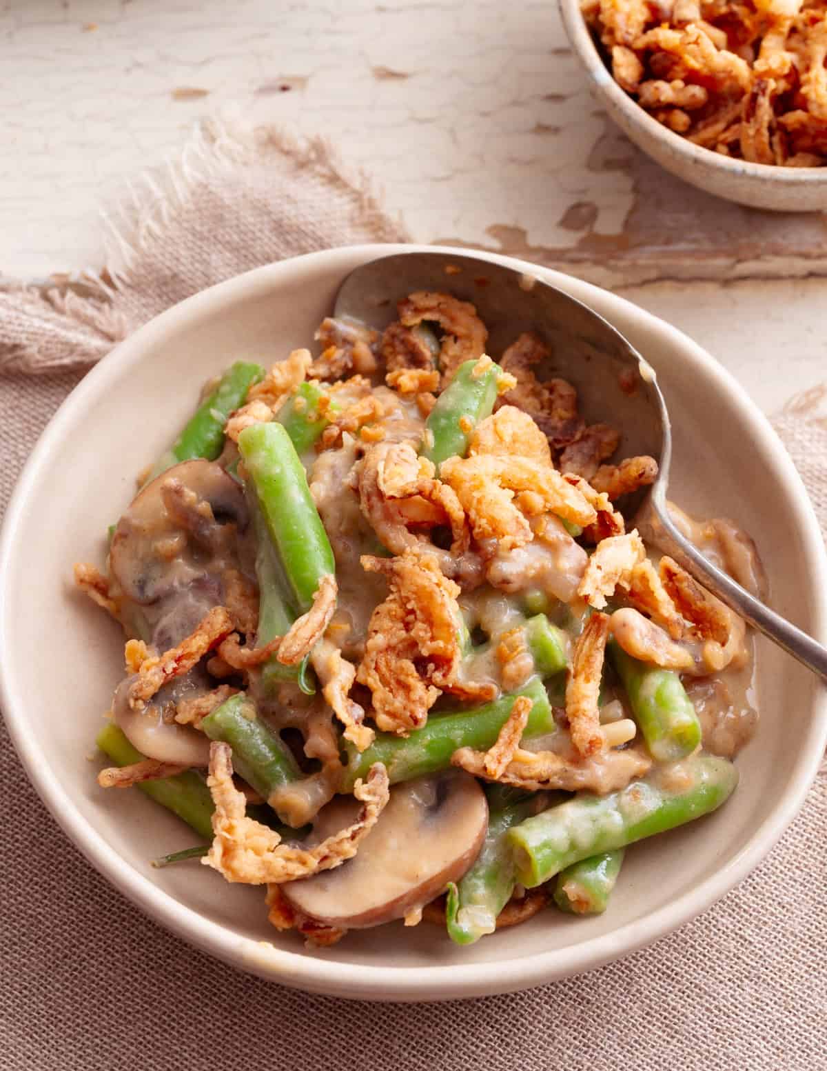 A small portion of vegan green bean casserole being spooned into a cream coloured bowl sat on a aged white wooden table. 