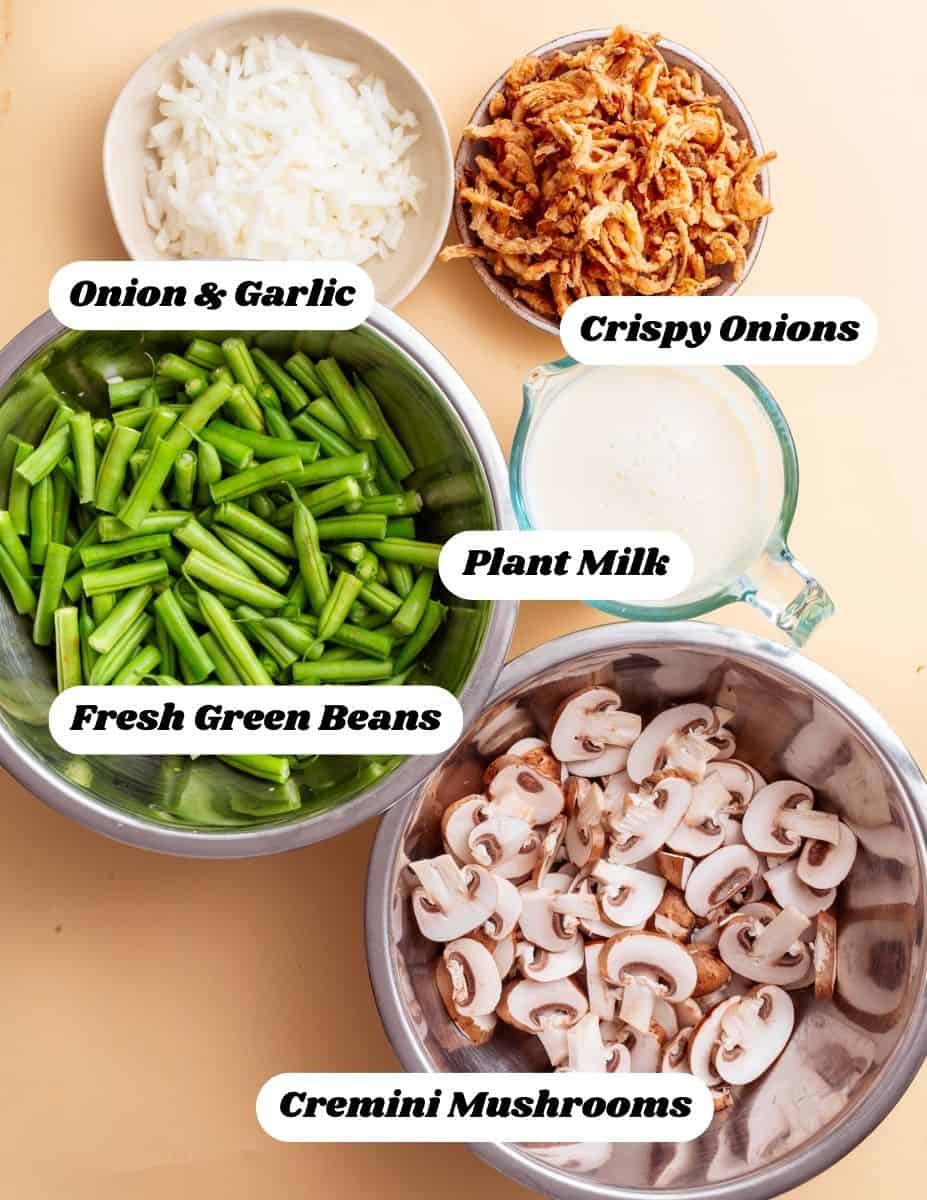 A top-down view of the main ingredients required for this recipe. In bowls there are onion and garlic, crispy onions, plant milk, fresh green beans and cremini mushrooms.