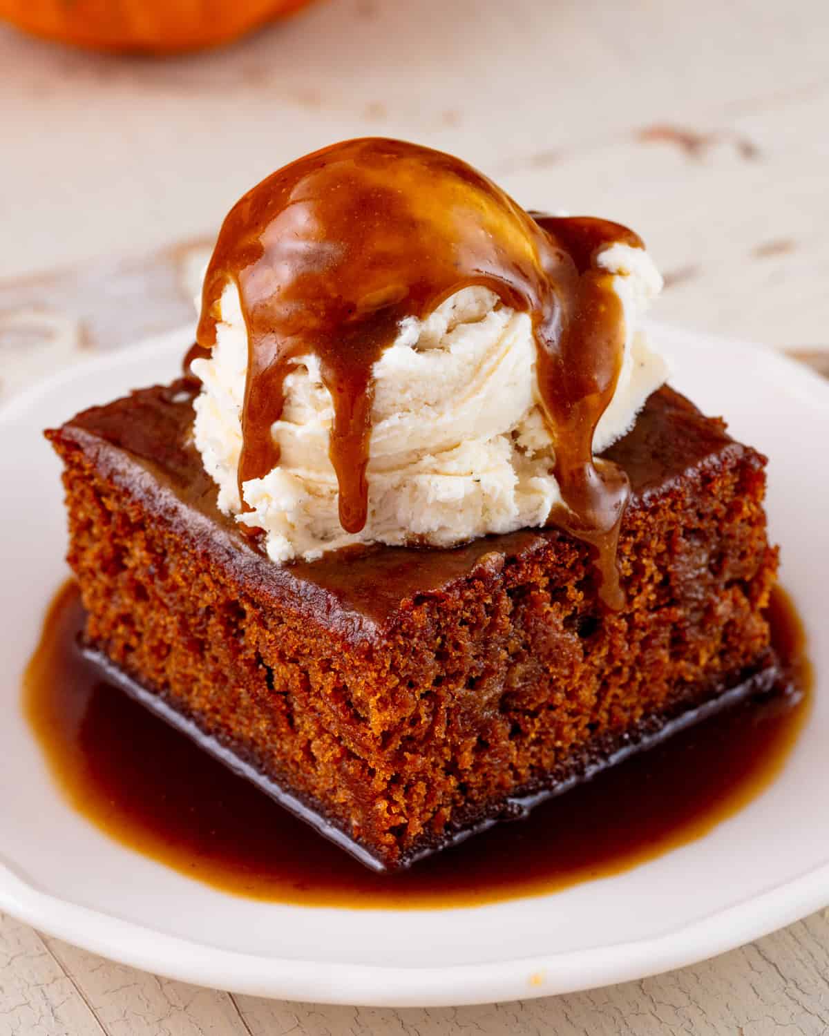 A slice of warm vegan pumpkin sticky toffee pudding on a plate with a scoop of ice cream and topped with hot toffee sauce.