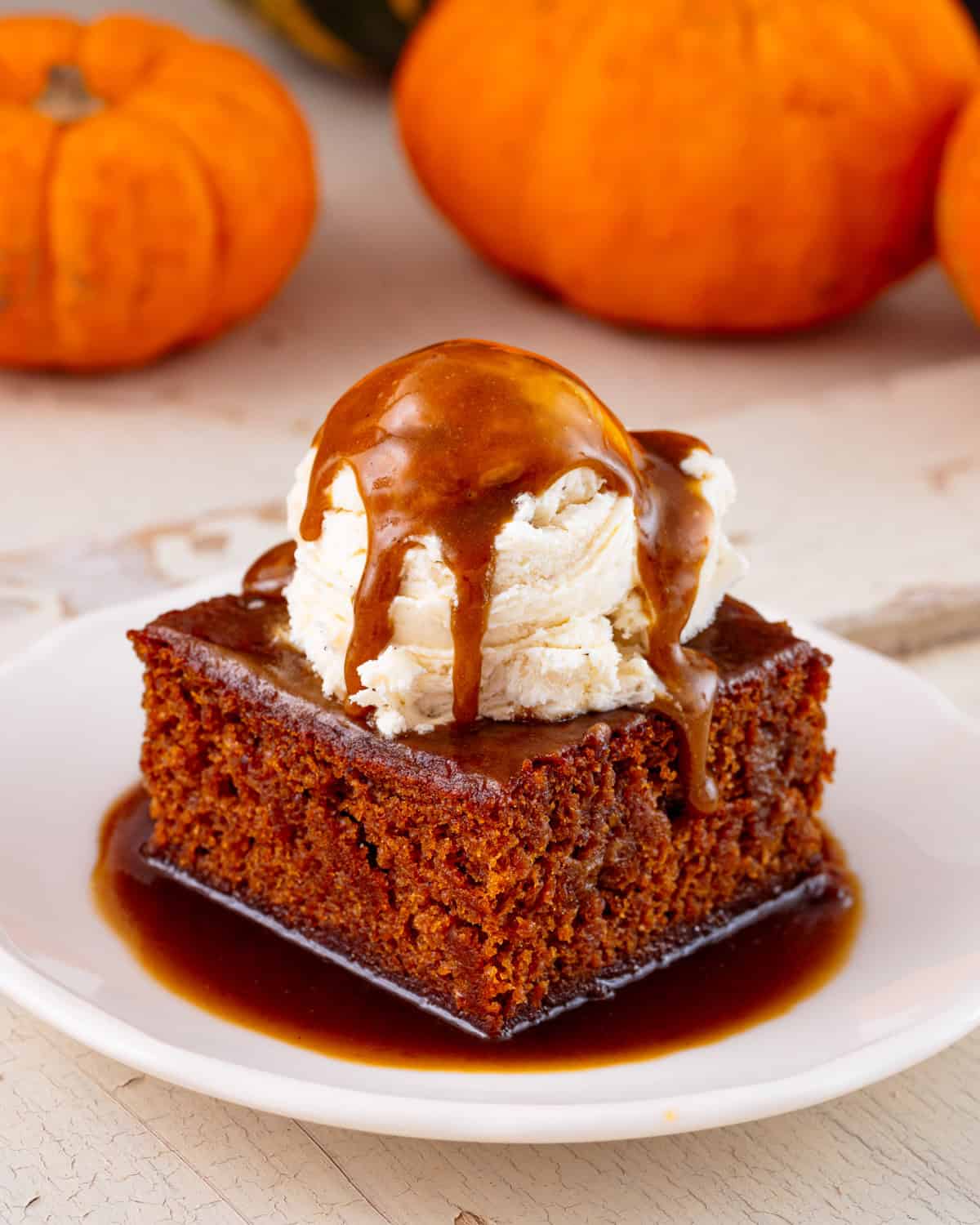 A slice of vegan pumpkin sticky toffee  pudding on a white plate with ice cream and toffee sauce on top.