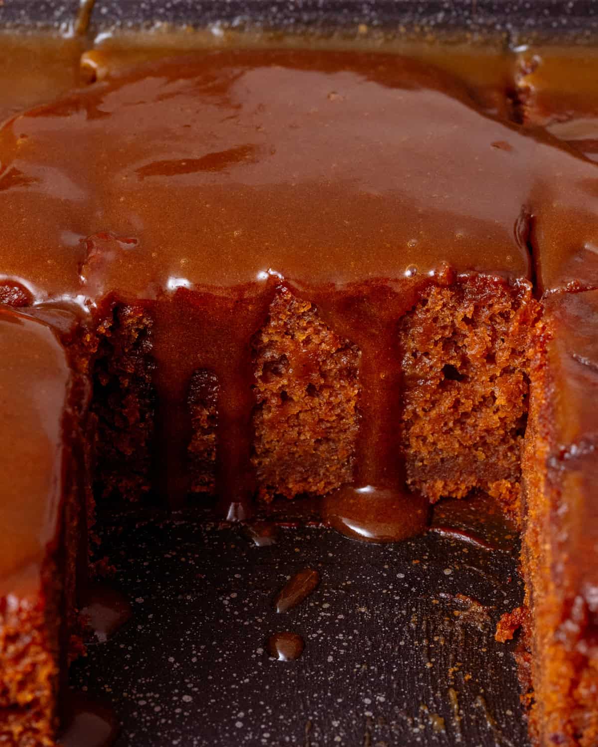 a slice of vegan sticky toffee pudding topped with pumpkin toffee sauce in a black brownie tin.