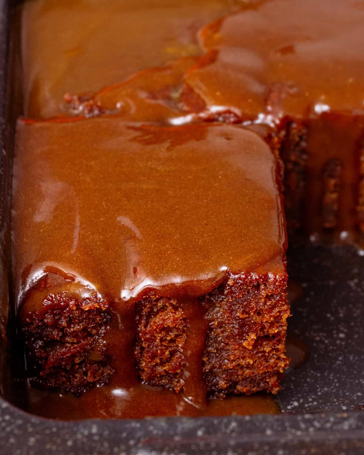 a slice of vegan sticky toffee pudding topped with pumpkin toffee sauce in a black brownie tin.