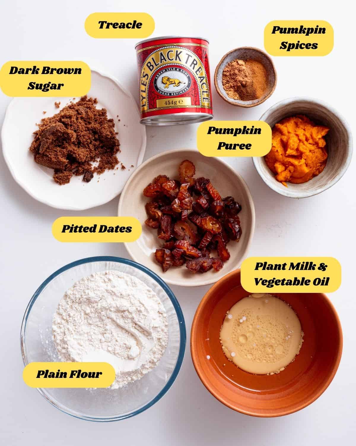 Ingredients you'll need for this recipe on a flat surface in bowls. This includes pitted dates, pumpkin puree, black treacle, plain flour, plant milk, vegetable oil, dark brown sugar and pumpkin spices.