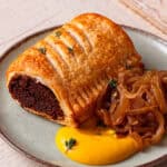A vegan sausage roll on a small plate served with mustard and caramelised onions with sprigs of thyme on top.