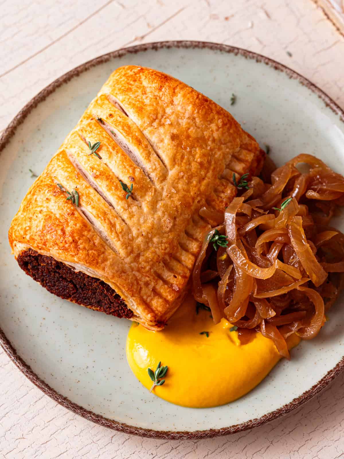 A vegan sausage roll with golden brown puff pastry on a small plate served with caramelised onions and mustard.