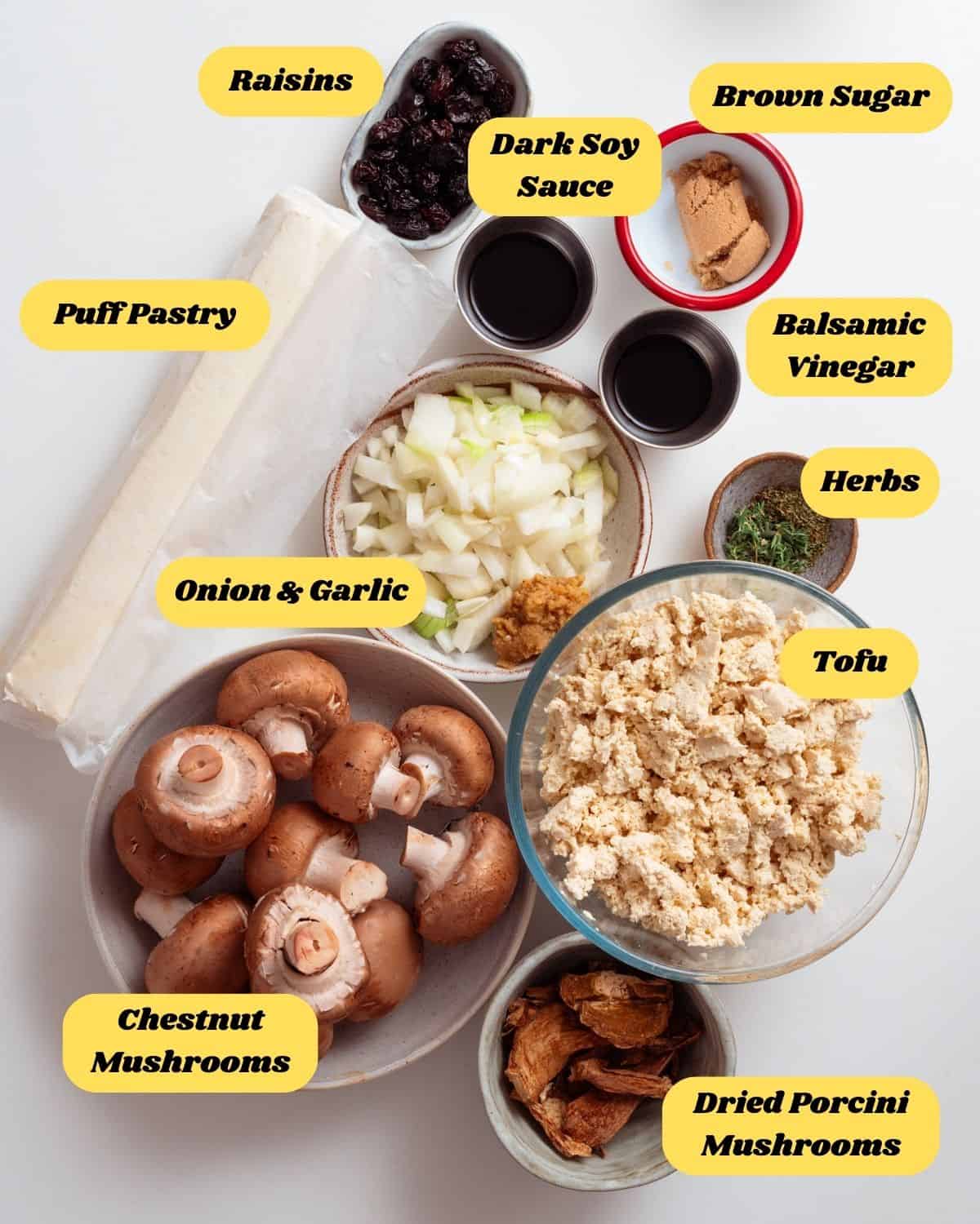 All the main ingredients required for this recipe, on a white table with their names listed. Including raisins, dark soy sauce, brown sugar, puff pastry, onion & garlic, balsamic vinegar, herbs, tofu, chestnut mushrooms, dried porcini mushrooms.